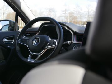 Binnenaanzicht van een Renault Captur-auto, gericht op het stuur met het Renault-logo, met zichtbaar dashboard en bestuurdersdeur.