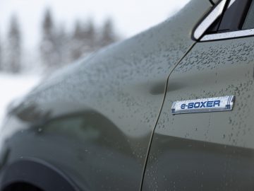 Close-up van het embleem van een Subaru Forester e-boxer met waterdruppels op de carrosserie, wat duidt op hybride technologie in een winterse omgeving.