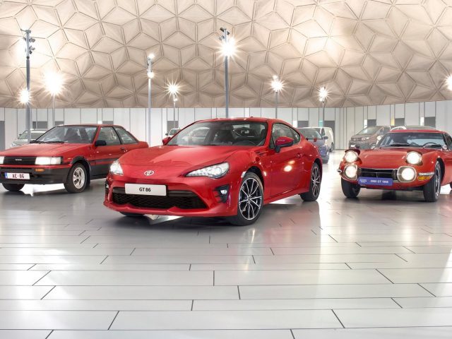 Rode Toyota GT86-sportwagen tentoongesteld naast twee klassieke modellen in een overdekte tentoonstellingsruimte.