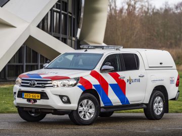 Een Nederlandse politieauto van Toyota staat buiten geparkeerd met een moderne bouwconstructie op de achtergrond.