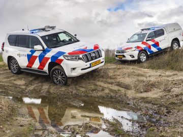 Twee Nederlandse politievoertuigen, waaronder een Toyota, op ruw terrein onder bewolkte hemel.