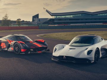Twee krachtige sportwagens, waaronder een Aston Martin, op een racecircuit, met moderne architectuur op de achtergrond.