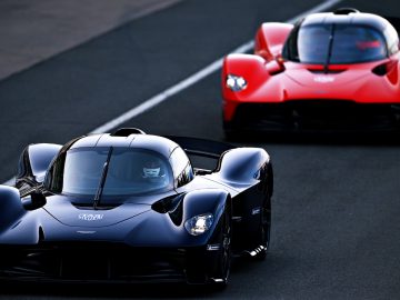 Twee krachtige sportwagens op het circuit, de ene zwart op de voorgrond en de andere rood erachter, beide Aston Martins.