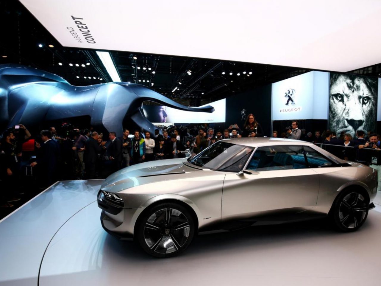 Bezoekers van Autosalon Parijs observeren een strakke concept-car met dynamische sculpturen en branding op de achtergrond.