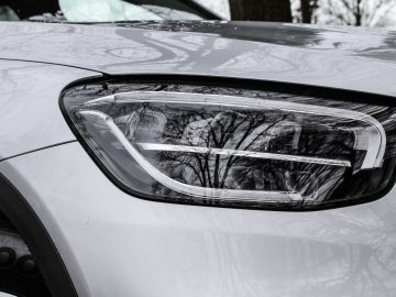 Close-up van de koplamp van een Mercedes-Benz GLC met een weerspiegeling van kale bomen op de motorkap.