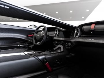 Binnenaanzicht van een Aston Martin V12 Speedster met koolstofvezeldetails, een stuur met logo, digitale displays en rode veiligheidsgordels.