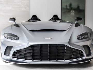 Vooraanzicht van een moderne zilveren Aston Martin V12 Speedster met een grote zwarte grille en een strak ontwerp, weergegeven in een minimalistisch interieur.