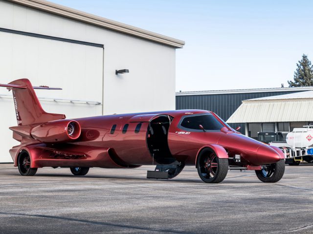 Een rode, op een vliegtuig geïnspireerde limousine met straalmotoren geparkeerd buiten een hangar.