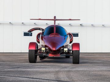 Een rode limo-jet-gyrocopter met open zijdeuren geparkeerd op asfalt met een hangar op de achtergrond.