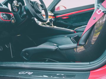 Binnenaanzicht van een Porsche 718 Cayman GT4-sportwagen, met de nadruk op de bestuurdersstoel, het stuur en de rode veiligheidsgordel.