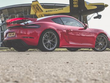 Rode Porsche 718 Cayman GT4 sportwagen geparkeerd op asfalt met een tweedekker van het merk Breitling op de achtergrond.