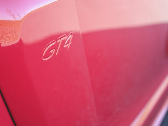 Close-up van een rode Porsche 718 Cayman GT4 met een "gta"-embleem op het zijpaneel.