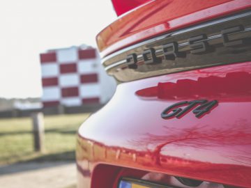 Close-up van het achterembleem van een rode Porsche 718 Cayman GT4 met een wazig gebouw op de achtergrond.