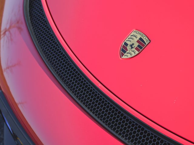 Close-up van een Porsche 718 Cayman GT4-embleem op de motorkap van een rode auto met daaronder een honingraatgrillepatroon.