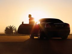 Man leunt op een Autonamen-luxeauto terwijl hij naar de zonsondergang kijkt op een open weg.