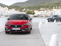 Een rode Seat Leon die over een kustweg rijdt met witte gebouwen op de achtergrond.