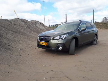 Een Subaru Outback geparkeerd op zanderig terrein met industriële constructies op de achtergrond.