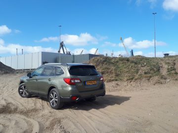Subaru Outback geparkeerd op zandig terrein met industriële constructies op de achtergrond.