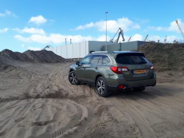 Groene Subaru Outback geparkeerd op zanderig terrein met industriële kranen op de achtergrond.