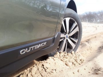 Een Subaru Outback-voertuig rijdt gedeeltelijk off-road, met zijn voorband op zandig terrein.