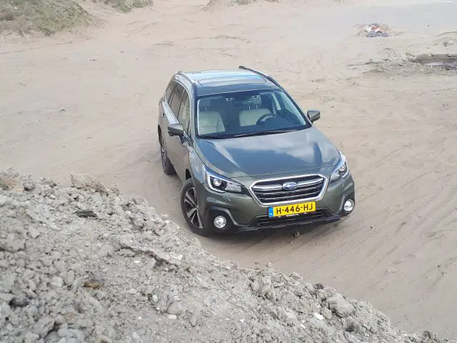 Een grijze Subaru Outback geparkeerd op een zandvlakte vlakbij een hoop aarde.