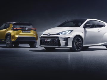 Twee Toyota Yaris Cross-modellen naast elkaar geparkeerd, een gele en een witte, tegen een donkere achtergrond.