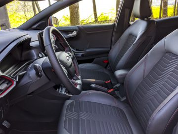 Binnenaanzicht van een moderne Ford Puma met de bestuurdersstoel, het stuur en het dashboard met zwarte bekleding en rode stiksels.