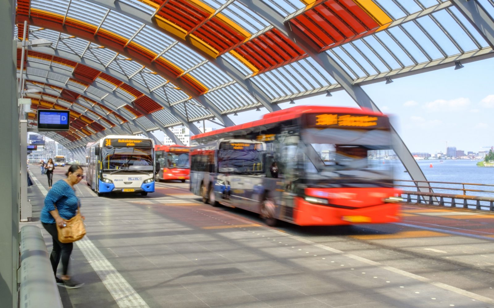 Bussen die op een zonnige dag aankomen en vertrekken bij een modern, overdekt busstation met een gebogen rood dak als onderdeel van het openbaar vervoersysteem.