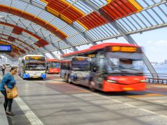 Bussen die op een zonnige dag aankomen en vertrekken bij een modern, overdekt busstation met een gebogen rood dak als onderdeel van het openbaar vervoersysteem.