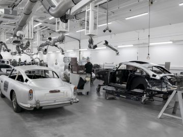 Moderne Aston Martin DB5 Goldfinger Vervolg restauratiewerkplaats met meerdere voertuigen in verschillende stadia van montage en een technicus die op de achtergrond werkt.