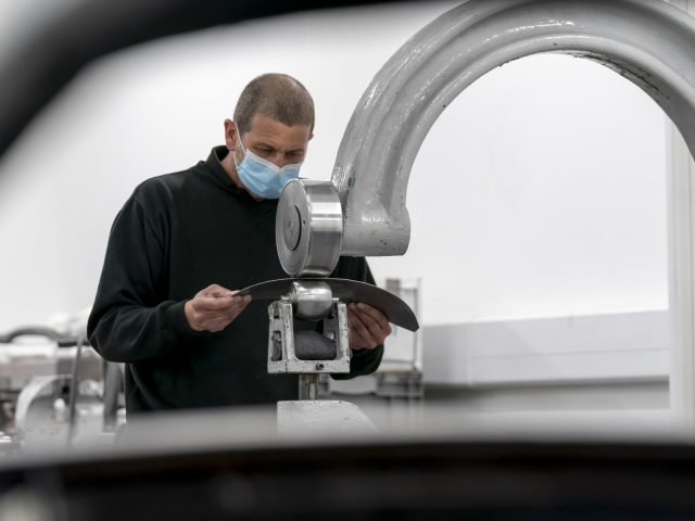 Een man met masker inspecteert een Aston Martin DB5 Goldfinger Continuation-onderdeel in een industriële omgeving.