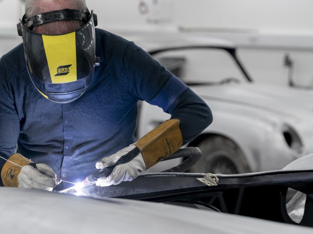 Een lasser die beschermende kleding draagt, is een metalen stuk aan het lassen in een werkplaats met een Aston Martin DB5 Goldfinger Continuation op de achtergrond.