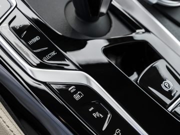 Close-up van de middenconsole van een BMW 5 Serie-auto met knoppen voor rijmodi, waaronder sport, hybride en elektrisch, evenals andere bedieningselementen van het voertuig.