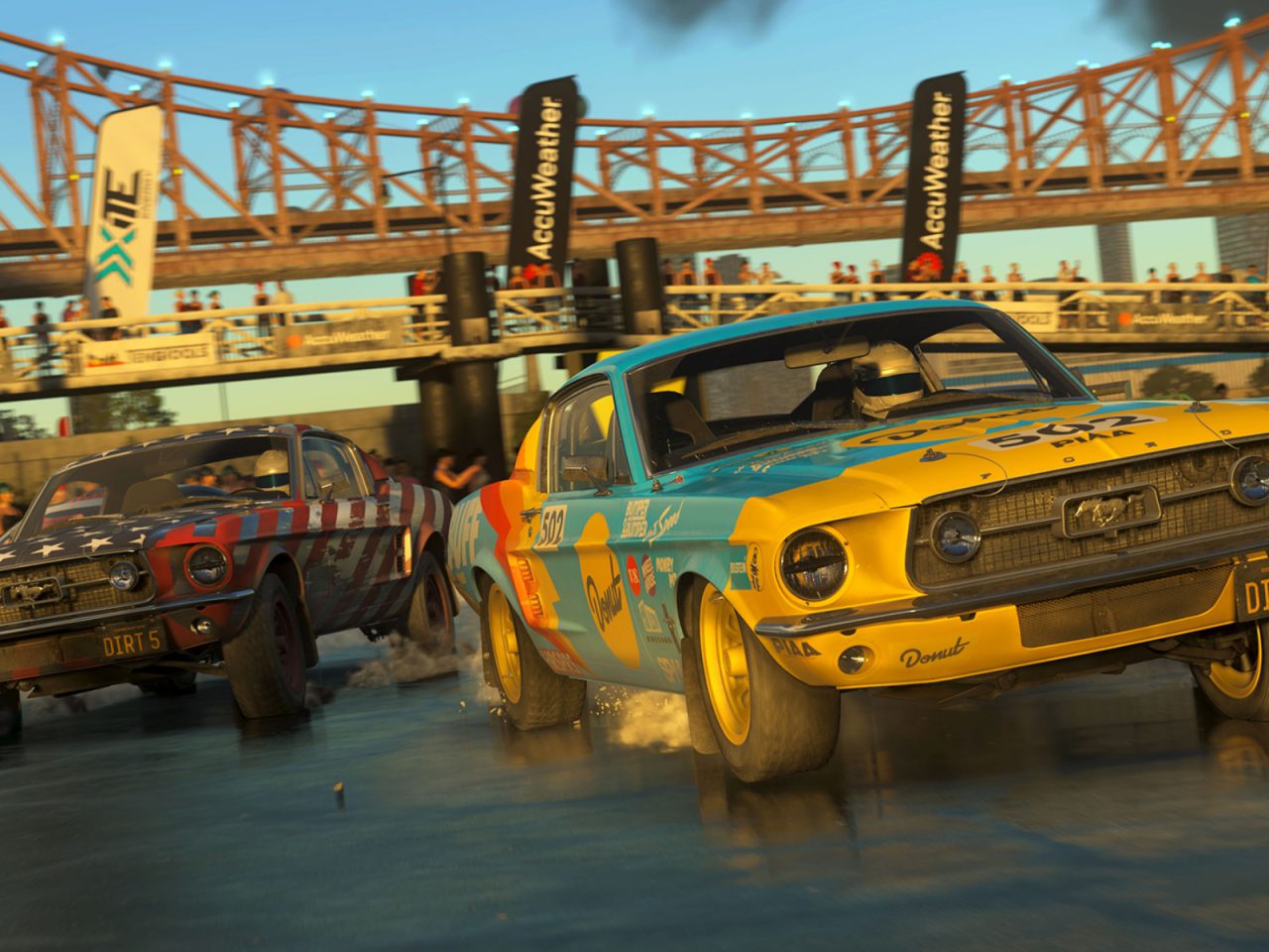 Twee vintage muscle car's racen op een onverharde weg bij zonsondergang in de DiRT 5-trailer, terwijl een menigte vanaf de zijlijn toekijkt.