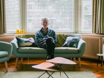 Een persoon die op een bank zit in een goed verlichte kamer met grote ramen en blinden, en leest over stikstof.