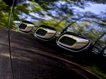 Een close-up van de glimmende zijkant van een Maserati Levante Trofeo, reflecterende bomen, met een rij stijlvolle uitlaatpijpen.