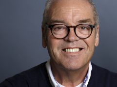 Close-up van een glimlachende man van middelbare leeftijd met bril tegen een F1 2020-achtergrond.