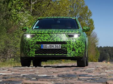 Een Opel Mokka met een groene camouflagewrap rijdt op een verharde weg met bomen op de achtergrond.