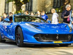 Een blauwe Ferrari-sportwagen rijdt door een straat met toeschouwers op de achtergrond tijdens een auto-evenement.