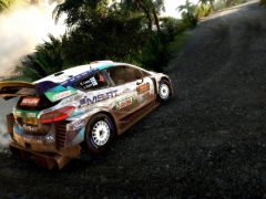 Een WRC 9-gameplay-rallyauto bedekt met modder concurreert op een onverharde weg omgeven door weelderige vegetatie.