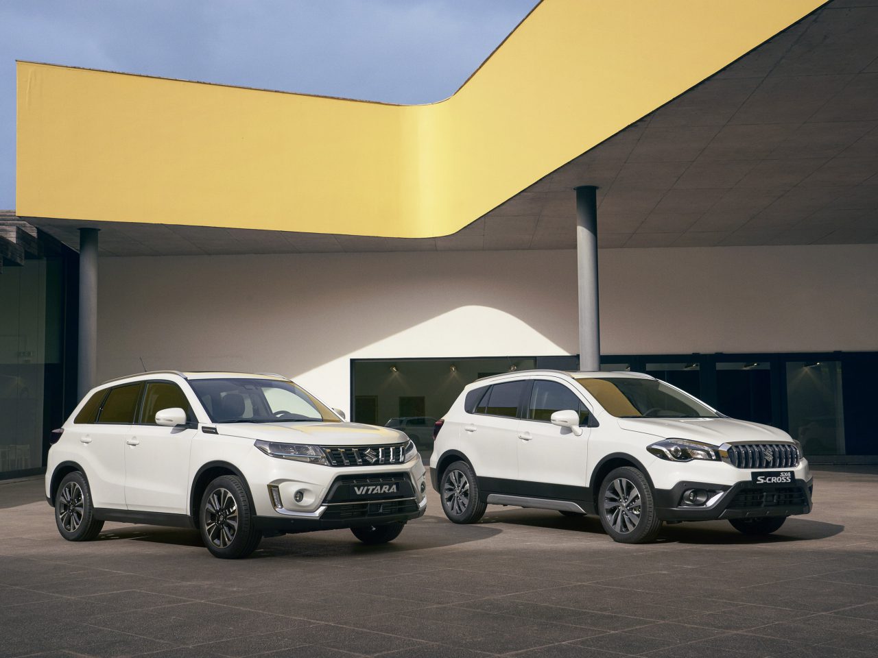 Twee Suzuki Vitara Smart Hybrid compacte SUV's staan naast elkaar geparkeerd voor een modern gebouw met een gele overhang.