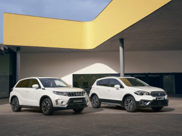 Twee Suzuki Vitara Smart Hybrid compacte SUV's staan naast elkaar geparkeerd voor een modern gebouw met een gele overhang.