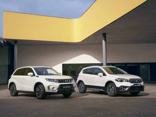 Twee Suzuki Vitara Smart Hybrid compacte SUV's staan naast elkaar geparkeerd voor een modern gebouw met een gele overhang.