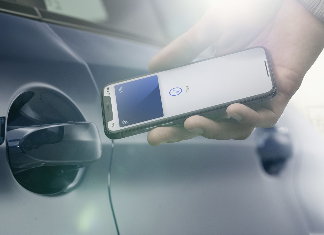 Hand die een iPhone vasthoudt met een oplaadsymbool op het scherm, uitgelijnd met de oplaadpoort van een elektrische auto van BMW, wat draadloze of contactloze oplaadtechnologie suggereert.