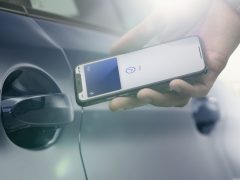 Hand die een iPhone vasthoudt met een oplaadsymbool op het scherm, uitgelijnd met de oplaadpoort van een elektrische auto van BMW, wat draadloze of contactloze oplaadtechnologie suggereert.