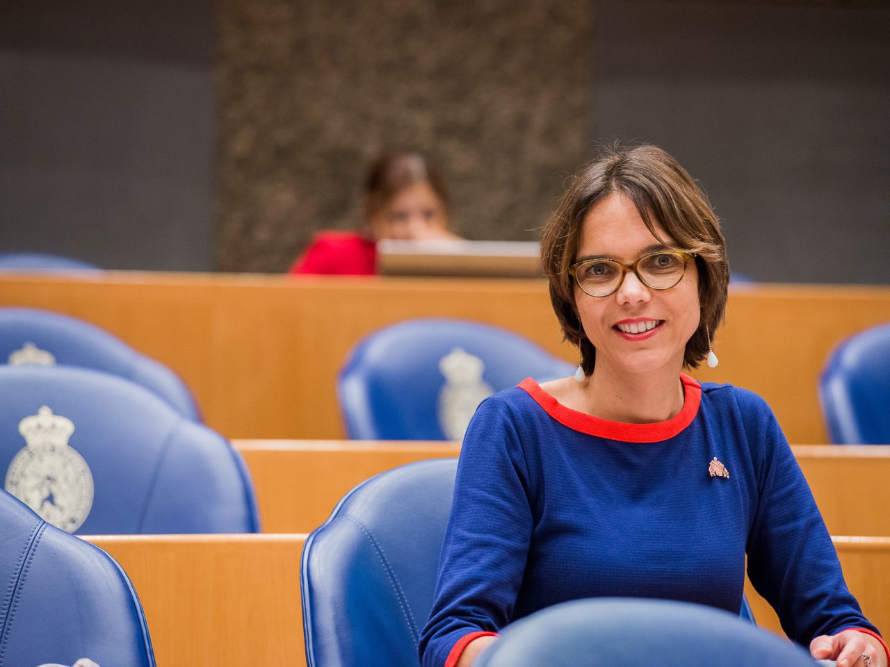 Een vrouw in een blauwe en rode jurk zit in een wetgevende vergaderzaal in Nederland.