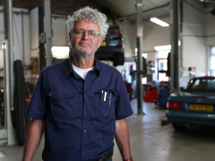 Monteur staat zelfverzekerd in een autoreparatiewerkplaats met een auto met verbrandingsmotoren op de achtergrond.