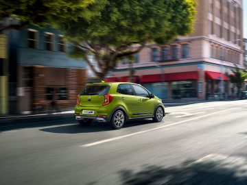 Een groene Kia Picanto die door een zonnige stadsstraat rijdt.