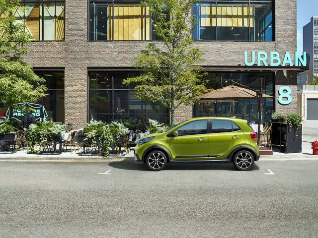 Heldergroene Kia Picanto geparkeerd in een zonnige straat naast een terras met moderne stedelijke architectuur op de achtergrond.