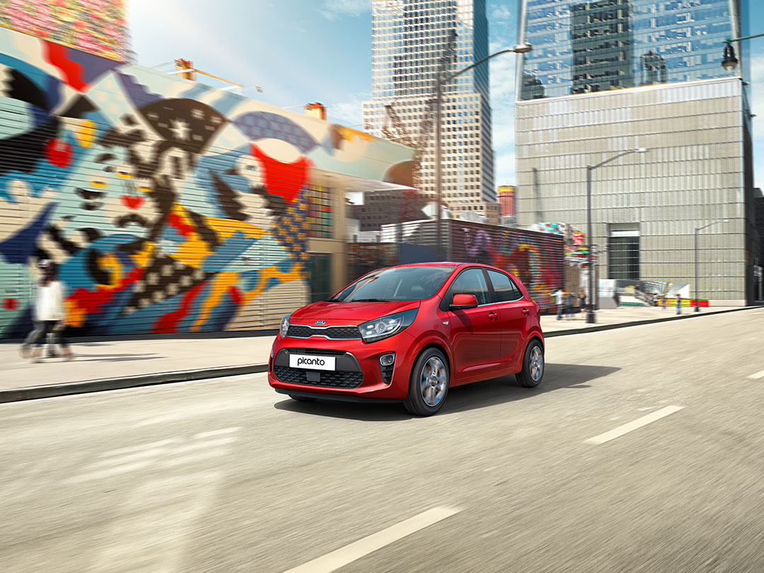 Rode Kia Picanto rijdt door een stedelijk gebied met kleurrijke straatkunst op de achtergrond.